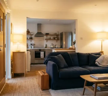 Moderne ingerichte woonkamer met comfortabele bank, keuken op achtergrond en koffer bij ingang, warm gouden licht op witte muren en eiken meubels.